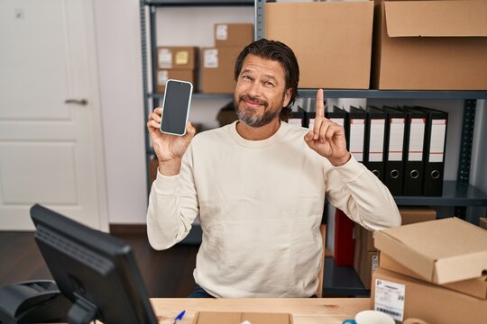 Handsome middle age man working at small business ecommerce holding smartphone surprised with an idea or question pointing finger with happy face, number one