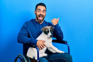Handsome hispanic man with beard sitting on wheelchair with puppy dog pointing thumb up to the side smiling happy with open mouth