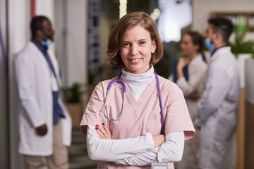 Young happy female general practitioner or nurse in medical scrubs crossing arms by chest while standing against intercultural colleagues