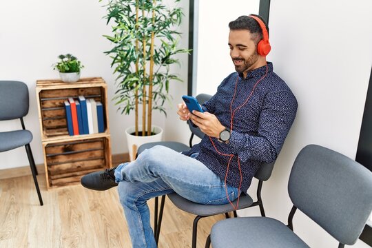 Young hispanic man listening to music sitting on chair at waiting room