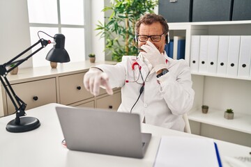 Senior doctor man working on online appointment laughing at you, pointing finger to the camera with hand over mouth, shame expression