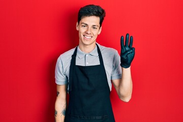 Young hispanic man wearing apron showing and pointing up with fingers number three while smiling confident and happy.