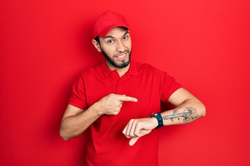 Hispanic man with beard wearing delivery uniform and cap in hurry pointing to watch time, impatience, upset and angry for deadline delay - Powered by Adobe
