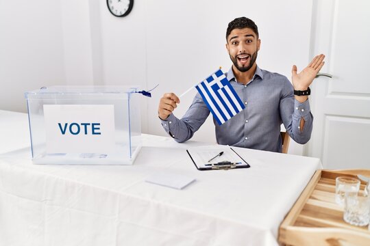 Young handsome man with beard at political campaign election holding greece flag celebrating victory with happy smile and winner expression with raised hands