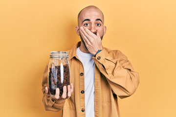 Young bald man holding jar of raisins covering mouth with hand, shocked and afraid for mistake. surprised expression