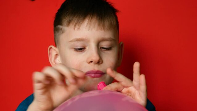Portrait of a cheerful caucasian boy 7 years old inflating a balloon on a red background. studio shooting. The child inflates balloons for the celebration. Festive mood, decoration with balloons