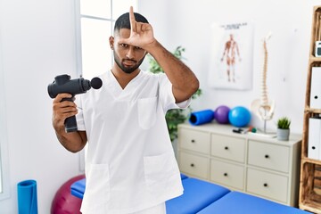 Young indian physiotherapist holding therapy massage gun at wellness center making fun of people with fingers on forehead doing loser gesture mocking and insulting.