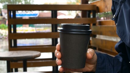 Close-up of a disposable paper cup for coffee is held by a woman hiding from the rain under the wooden veranda of a summer cafe in the city. Coffee invigorates and warms the body.
