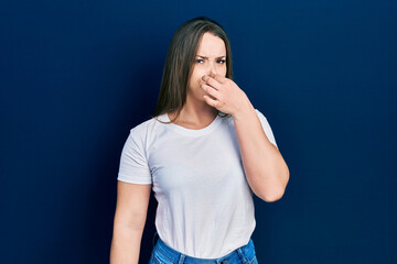 Young hispanic girl wearing casual white t shirt smelling something stinky and disgusting, intolerable smell, holding breath with fingers on nose. bad smell - obrazy, fototapety, plakaty