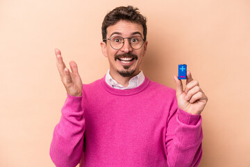 Young caucasian man holding batteries isolated on beige background receiving a pleasant surprise, excited and raising hands.