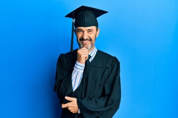 Middle age hispanic man wearing graduation cap and ceremony robe looking confident at the camera with smile with crossed arms and hand raised on chin. thinking positive.