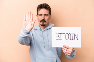Young caucasian man holding a bitcoin placard isolated on beige background standing with...