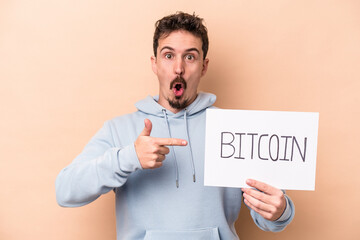 Young caucasian man holding a bitcoin placard isolated on beige background pointing to the side