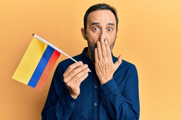 Middle age hispanic man holding colombia flag covering mouth with hand, shocked and afraid for...