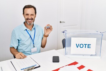 Middle age man with beard sitting by ballot holding i vote badge winking looking at the camera with...