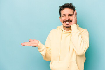 Young caucasian man isolated on blue background holds copy space on a palm, keep hand over cheek. Amazed and delighted.