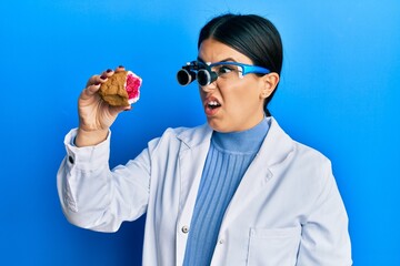 Beautiful brunette jeweller woman holding geode stone wearing magnifier glasses in shock face, looking skeptical and sarcastic, surprised with open mouth