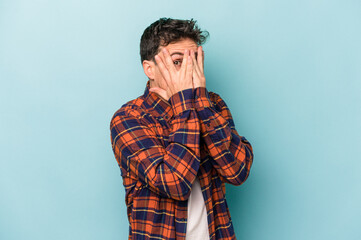 Young caucasian man isolated on blue background blink through fingers frightened and nervous.