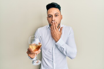 Young arab man drinking a glass of white wine covering mouth with hand, shocked and afraid for...