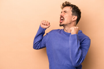 Young caucasian man isolated on beige background raising fist after a victory, winner concept.