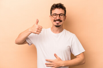 Young caucasian man isolated on beige background touches tummy, smiles gently, eating and satisfaction concept.