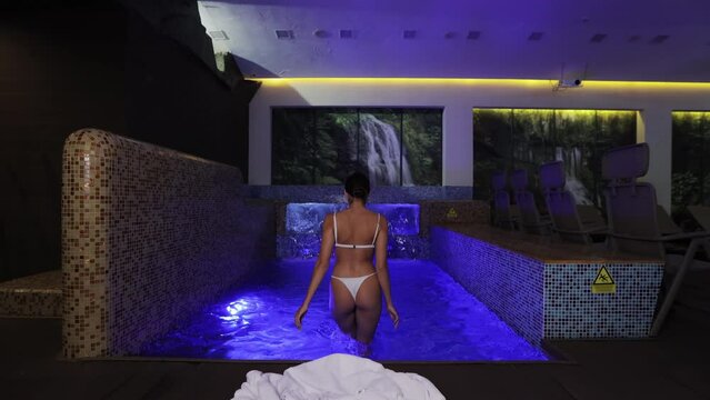 Back view of young woman with slender sexy body in white swimsuit swimming in pool at luxury hotel. Charming female person enjoying spa time during vacation.