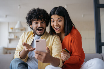 Overjoyed multiracial couple shopping online with big sales looking at digital screen sitting at home. Happy African American woman holding credit card Indian man using mobile phone ordering food