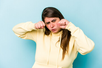 Young caucasian overweight woman isolated on blue background throwing a punch, anger, fighting due to an argument, boxing.