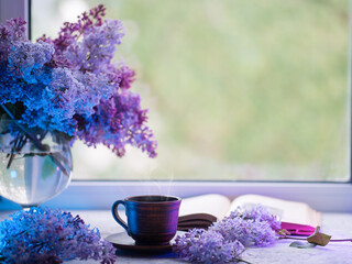 Spring still life with a cup of coffee and bouquets of lilacs. Colored light. Reading a book and drinking coffee by the window, spring mood