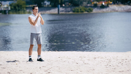 young sportsman in tank top and shorts warming up fingers near river