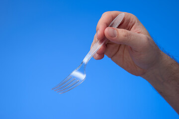 Transparent plastic single use fork held in hand by man. Close up studio shot, isolated on blue...