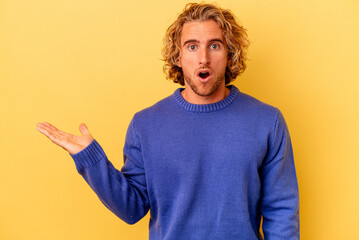 Young caucasian man isolated on yellow background impressed holding copy space on palm.