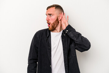 Young caucasian man isolated on white background trying to listening a gossip.