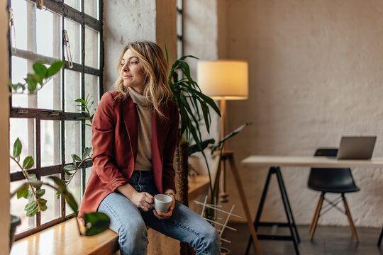 Thoughtful blonde woman, gazing outside the window, sitting and sipping tea. - Powered by Adobe