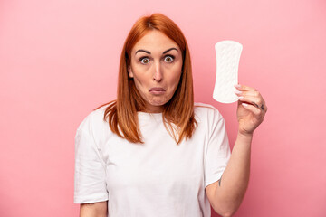 Young caucasian woman holding sanitary napkin isolated on pink background shrugs shoulders and open eyes confused.