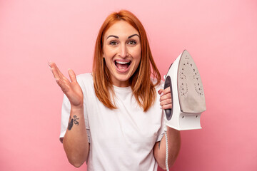 Young caucasian woman holding iron isolated on pink background receiving a pleasant surprise,...