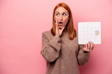 Young caucasian woman holding puzzle isolated on pink background is saying a secret hot braking news and looking aside