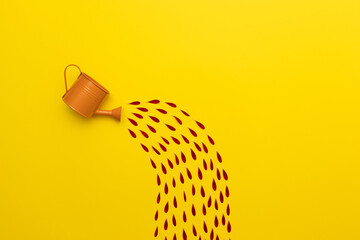 Orange watering can with abstract stream of water made of red petals on a yellow background, flat lay.