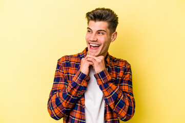 Young caucasian man isolated on yellow background keeps hands under chin, is looking happily aside.