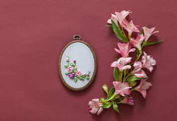 Spring needlework. Handmade gift for 8 March or birthday. Beautiful composition with alstroemeria flowers and floral embroidery with satin ribbons in hoop on brown background. Flat lay, copy space