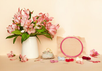 Spring still life with a bouquet of pink alstroemeria flowers, a canvas for cross-stitch embroidery in a hoop and skeins of thread for embroidery on a pale pink pastel background