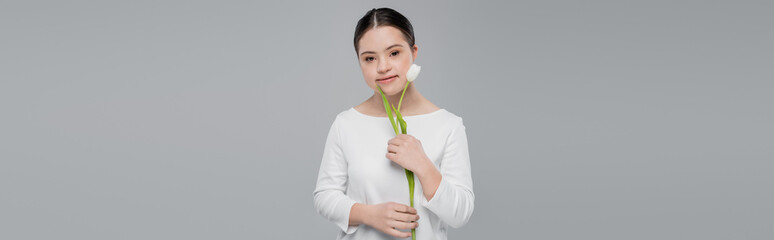Woman with down syndrome holding white tulip and looking at camera isolated on grey, banner