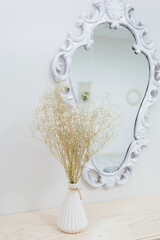 A bouquet of dry wildflowers stands in a white vase on a boudoir table by a vintage mirror.