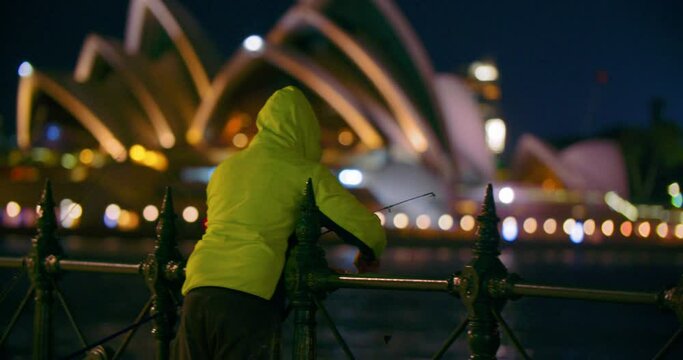 Rear View Of A Man In Yellow Hoodie Fishing In Darling Harbour At Night With Blurry Background Of Sydney Opera House In Australia.  - Selective Focus