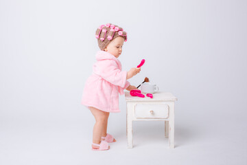 A cute little baby girl in a pink terry cloth and a curler robe, a mini dressing table and cosmetics is isolated on a white background, repeats the behavior of her mother