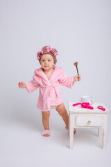 A cute little baby girl in a pink terry cloth and a curler robe, a mini dressing table and cosmetics is isolated on a white background, repeats the behavior of her mother