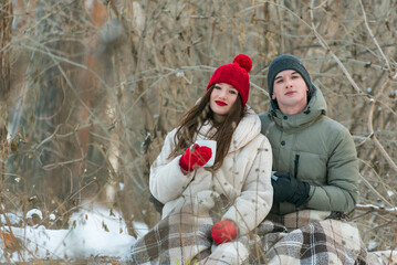 Portrait of young couple are sitting hugging each other in a snow-covered winter park, wrapping themselves in a plaid. Man and woman on a picnic in the winter forest.