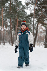 Fototapeta na wymiar Funny child stands in snowy forest and looking at the camera. Boy stands against of beautiful winter landscape