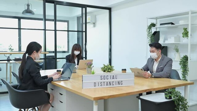 Group of young Asian team business people with face masks work in office and keep distancing in company workplace, Social Distancing concept
