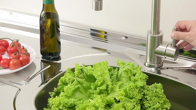 Hand opening a kitchen sink faucet and water spiling and poring over a lettuce, in slow motion.